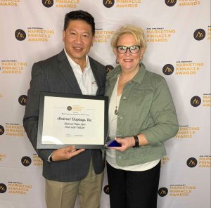 A man and a woman in business dress stand in front of a step and repeat backdrop holding awards