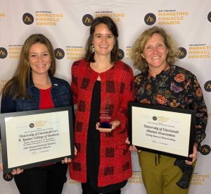 Three women stand in front of step and repeat backdrop holding awards and framed certificates